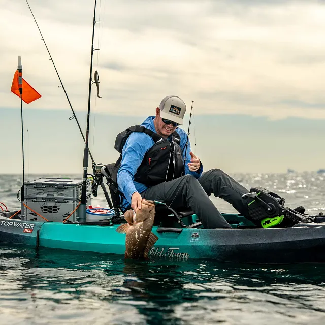 A teal and black Old Town fishing kayak floats on the water, equipped with a black seat, a black and green ePDL DRIVE pedal system, a gray storage crate, and fishing rods.