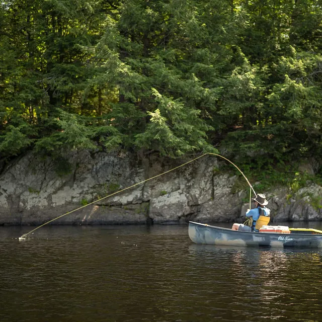 Old Town: An Old Town canoe, dark gray with camouflage patterns, floats on dark water with a person wearing a hat and yellow life vest seated inside, holding a fishing rod with a line cast into the water, a white cooler, and a yellow paddle, against a background of a rocky shoreline and dense green trees.