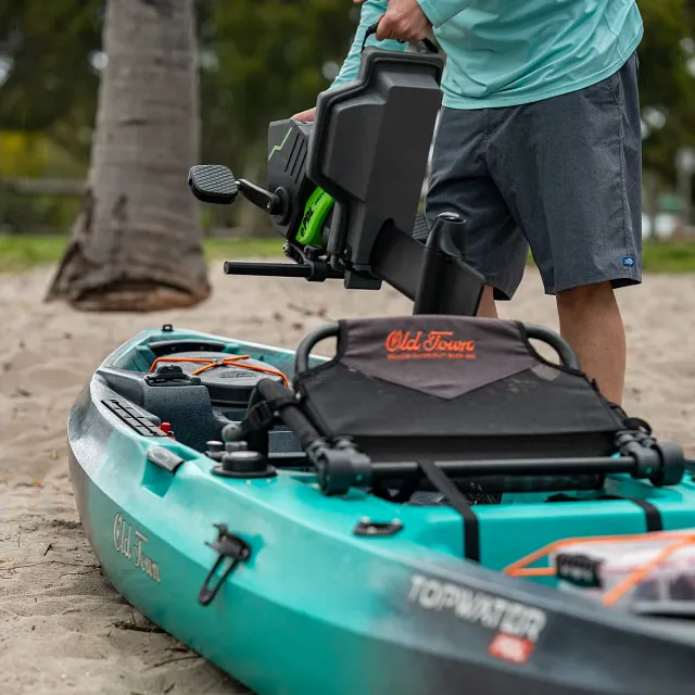 An Old Town Topwater kayak, teal and grey with a black fabric seat, orange bungee cords, and black rails, rests on a sandy beach while a person holds a black and green motor drive unit above it.