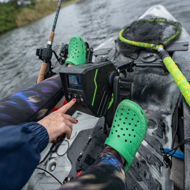 Old Town: A white Old Town kayak is on the water, with a black motor control head featuring a screen, buttons, and green accents mounted in the cockpit, where a person's hand presses a button.