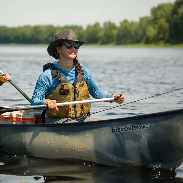 Old Town: An Old Town Discovery 158 canoe with a dark gray camouflage hull floats on calm water, with a person wearing a brown hat and tan life vest paddling while holding a fishing rod.