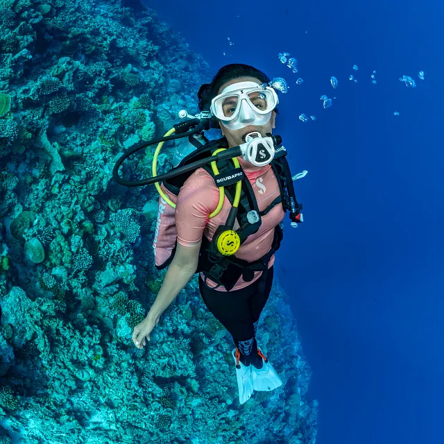 Plongeur sous‑marin portant un système de détendeur MK11 EVO INT/C370 de couleur blanche et un gilet stabilisateur Navigator Lite, nageant le long d’un récif corallien dans une eau bleu clair.