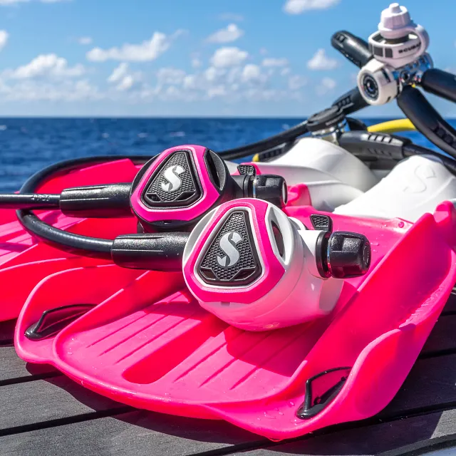 Zwarte en witte C370‑duikregelaars met roze kleurframe liggend op roze Seawing Supernova‑vinnen naast het water.