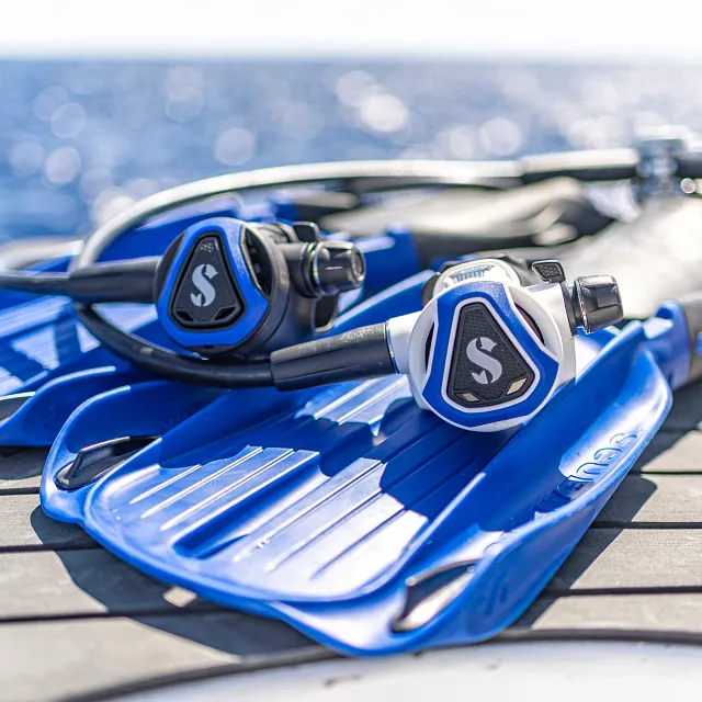 Schwarzer und weißer C370‑Tauchatemregler mit blauem Farbrahmen auf blauen Seawing‑Supernova‑Flossen neben dem Wasser.