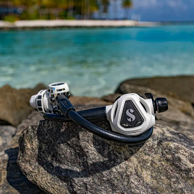 Weißer C370‑Tauchatemregler, der auf einem Felsen neben klarem blauem Wasser liegt.