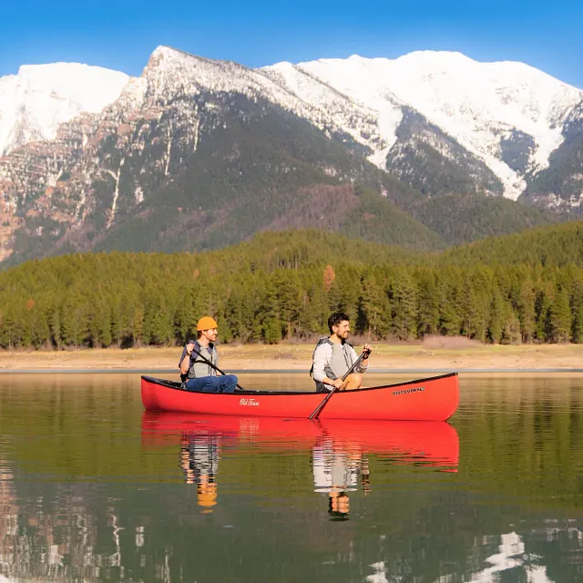 Old Town: A red canoe with black trim is being paddled by two men wearing grey life vests on a calm lake, with a forested shoreline and snow-capped mountains visible in the background under a clear blue sky.