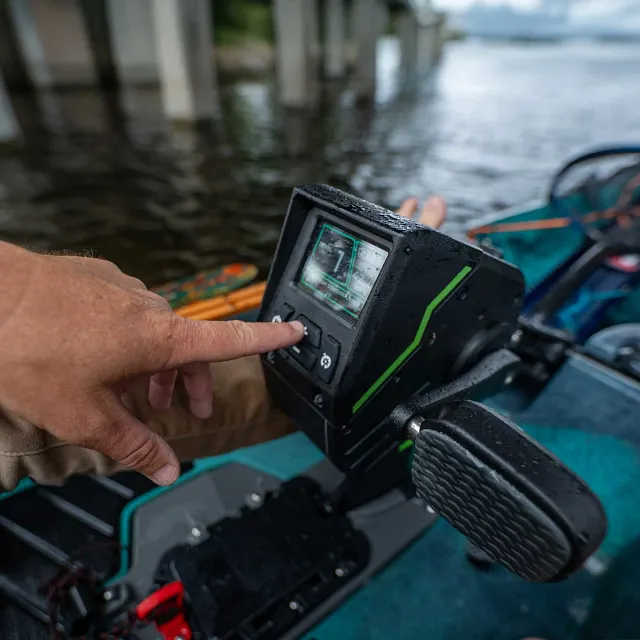 A black kayak motor control head with green accents, featuring a screen and several buttons, is mounted on a teal kayak, with a person's hand pressing a button.