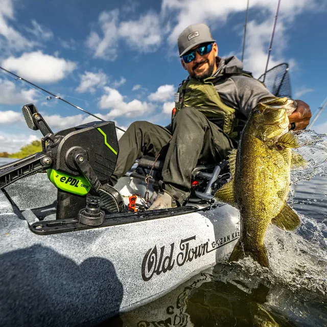 A light gray Old Town kayak with a black and lime green ePDL Drive unit is on the water, with a man in a gray hat and olive green life vest holding a large fish that splashes out of the water.