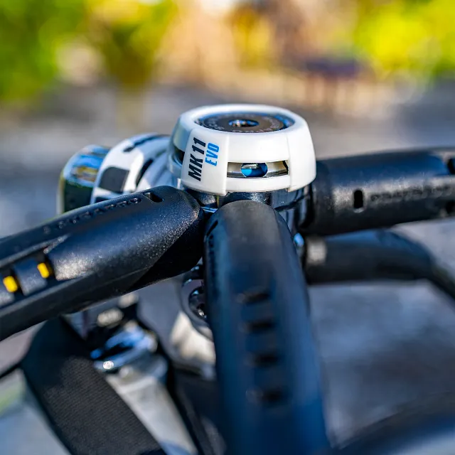 Gros plan du premier étage du détendeur de plongée MK11 EVO en blanc, avec des tuyaux raccordés, sur un fond extérieur.